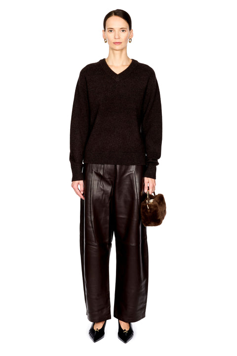 A model stands against a white background wearing a dark brown v-neck knit sweater, wide-leg dark brown faux-leather pants, black pointed shoes, and holding a small brown fur handbag.