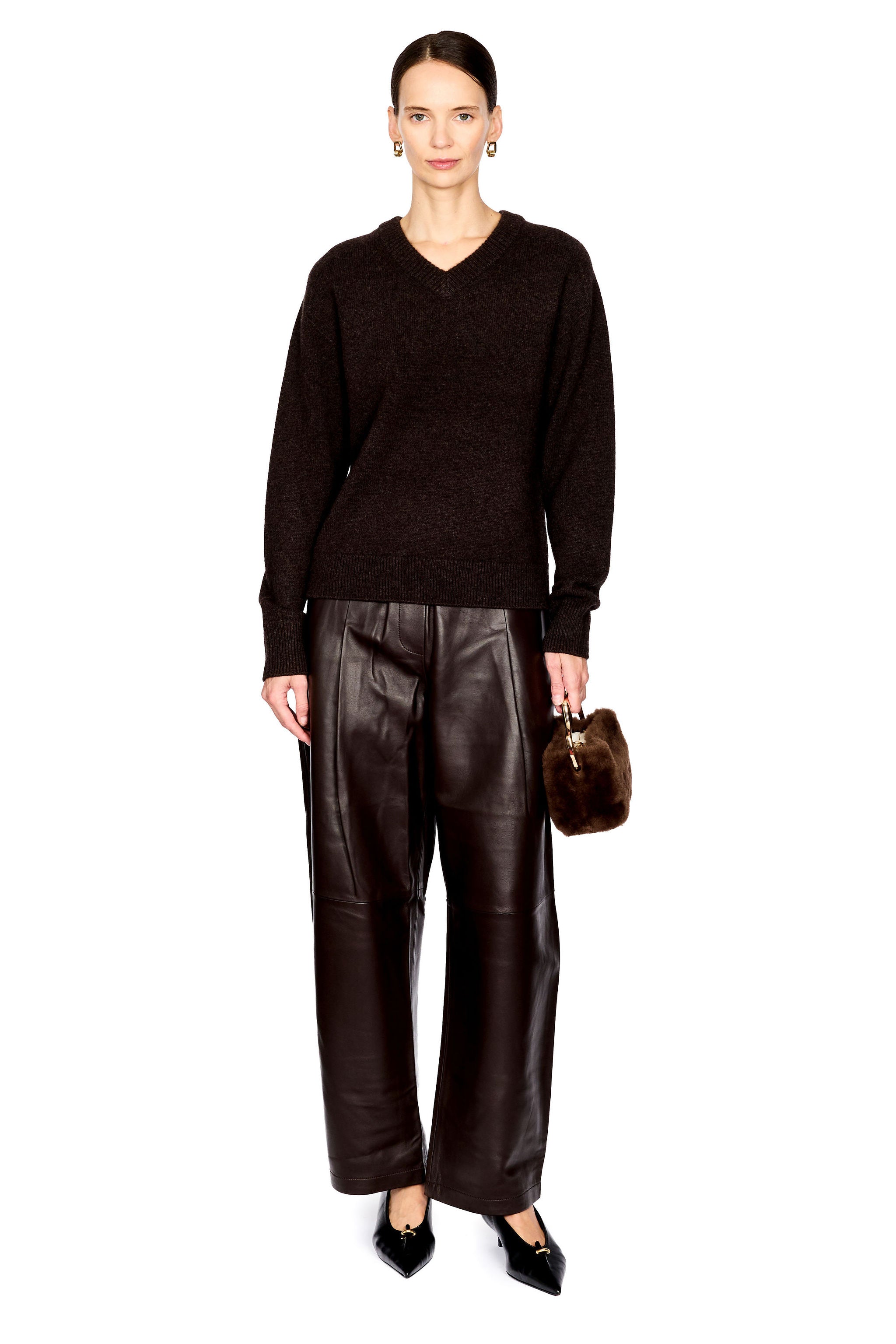 A model stands against a white studio background wearing a dark brown knit V-neck sweater, dark brown wide-leg leather pants, black pointed flats, and holding a small dark brown fur-like handbag.