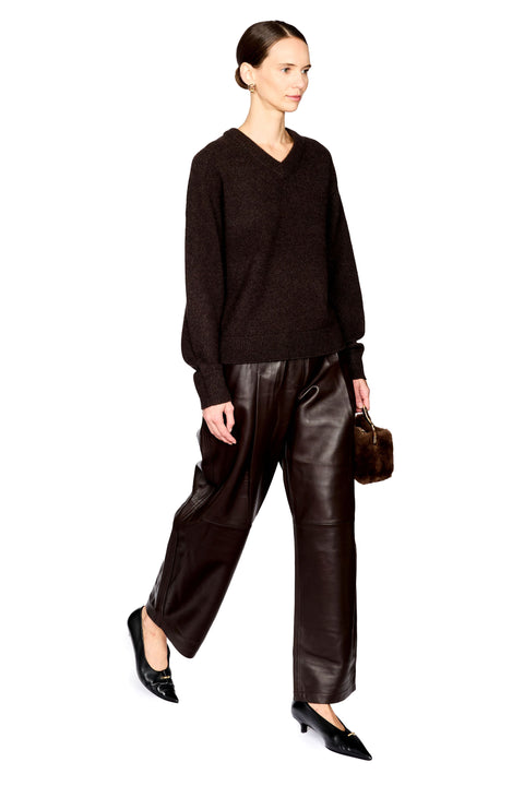 A model wearing a dark brown V-neck knit sweater, dark brown wide-leg leather pants, and black pointed flats, holding a small brown fur-trimmed handbag against a plain white background.