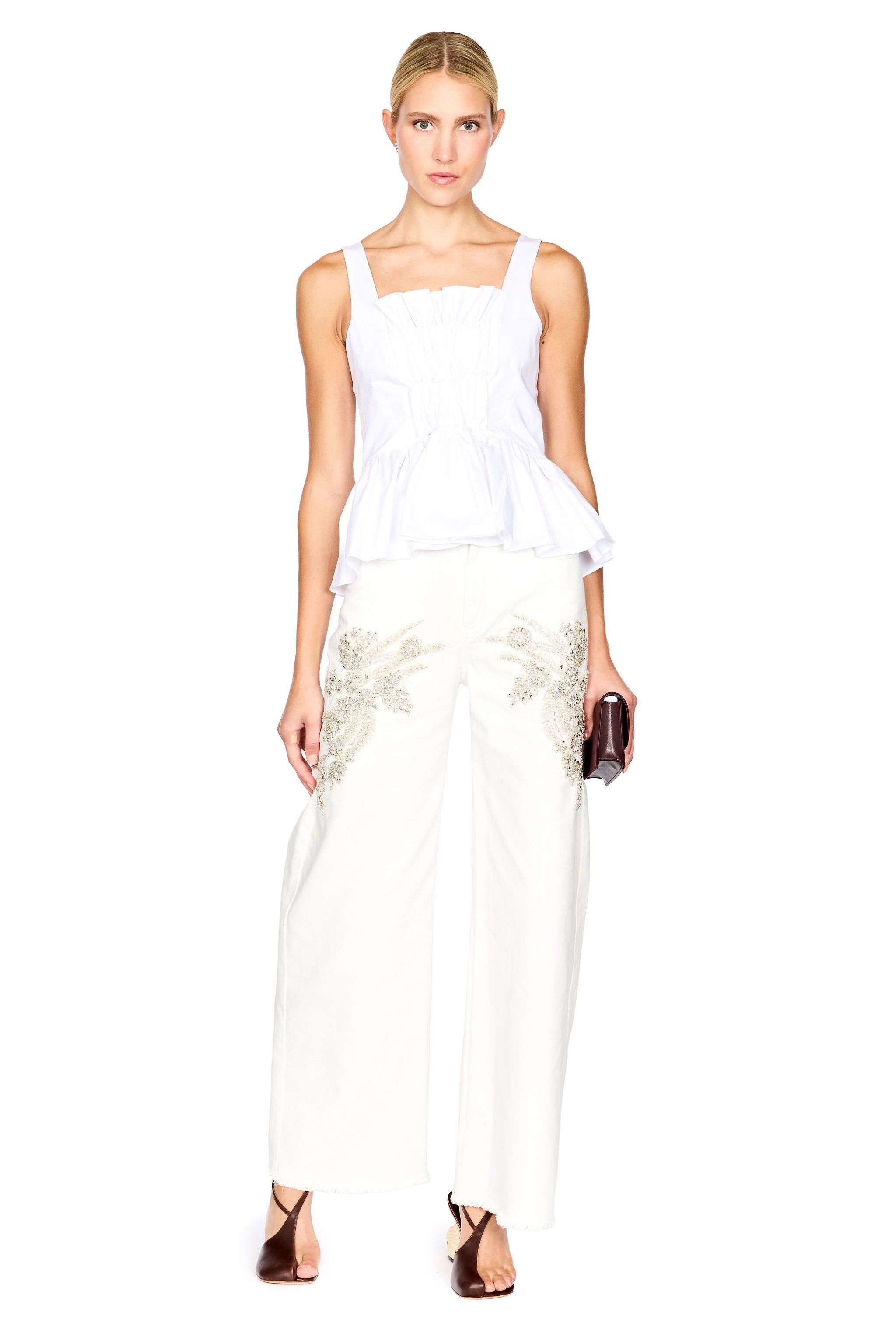 A woman modeling a white peplum top and white wide-leg trousers with gold floral embroidery, holding a small dark clutch against a plain white background.