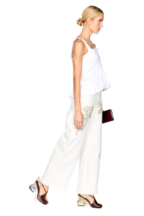 A blonde model in profile wearing a white sleeveless peplum top and white wide-leg trousers with gold embroidery, paired with brown heeled sandals and holding a small dark red clutch, set against a plain white background.