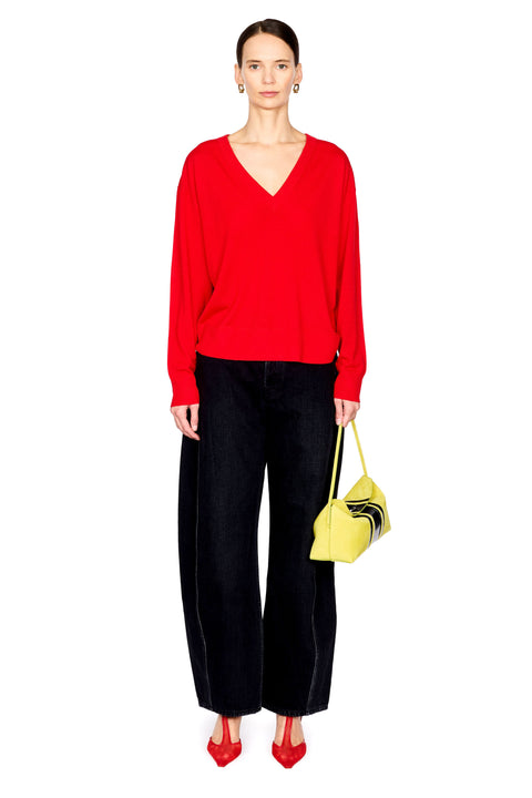 A model stands against a white studio background wearing a bright red long-sleeve V-neck sweater, black wide-leg trousers, red pointed heels, and holding a small yellow handbag.