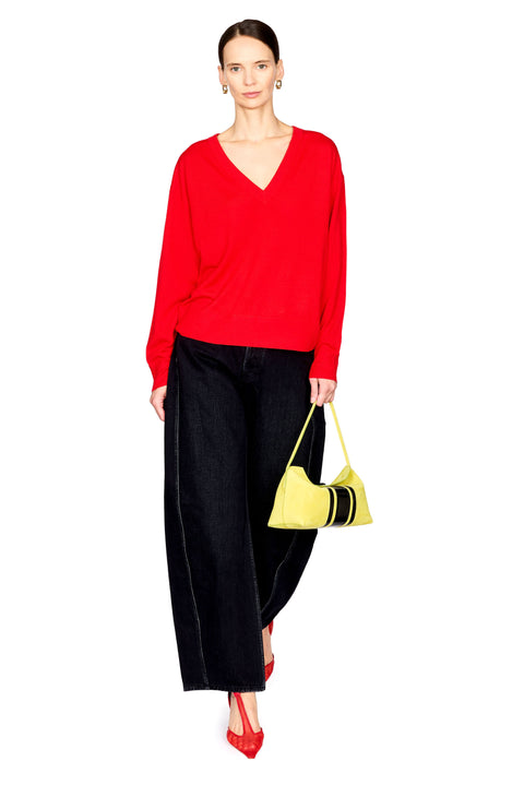 A model stands against a white background wearing a bright red long-sleeve V-neck sweater, black wide-leg trousers, red pointed heels, and holding a small yellow handbag with black stripes.