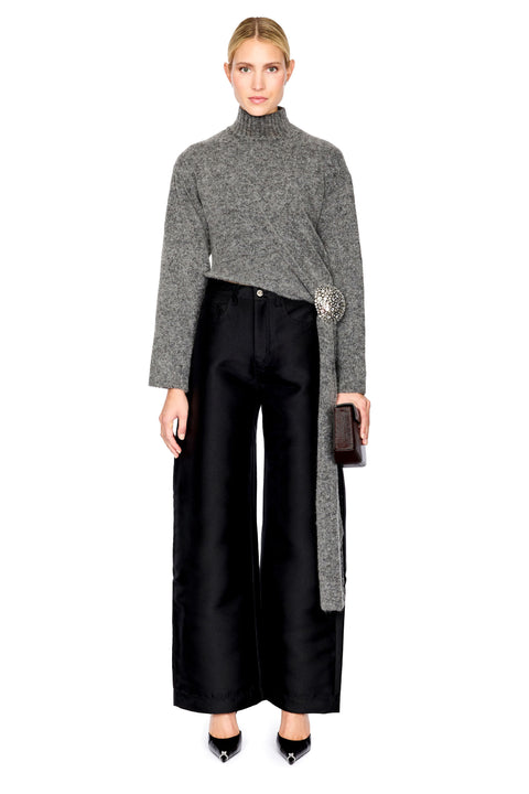 A model stands against a white background wearing a gray cropped turtleneck sweater with a long tie detail, black wide-leg trousers, and black pointed shoes, while holding a small black clutch.