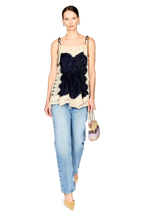 A model wearing a sleeveless camisole top with a navy velvet corset-style center and cream lace trim, paired with light blue jeans and holding a small striped handbag against a plain white background.