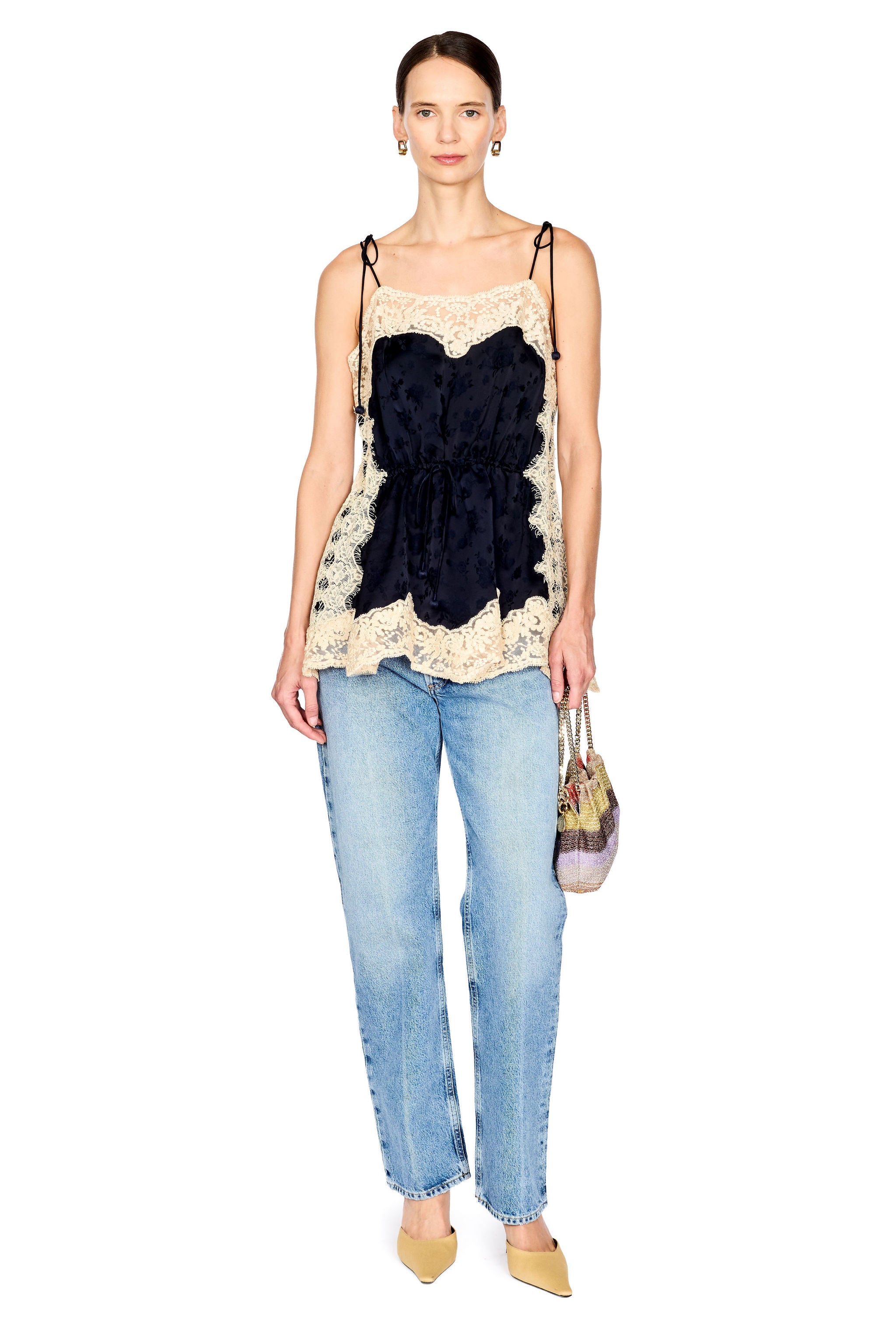 A model wearing a sleeveless camisole top with a large black ruffle motif over a cream lace border, paired with light-wash blue jeans and beige pointed shoes, holding a small patterned handbag against a plain white background.