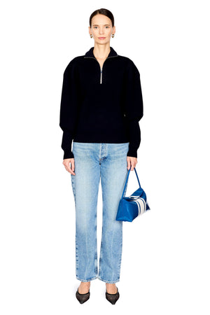 A model stands against a white background wearing a navy long-sleeve quarter-zip sweater, light-wash blue jeans, black pointed shoes, and holding a blue and white striped handbag.