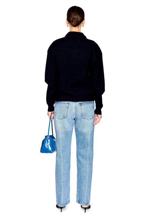 A woman modeling a navy long-sleeve quarter-zip sweater, light-wash blue jeans, and black pointed shoes, while holding a shiny blue handbag with white stripes against a plain white background.