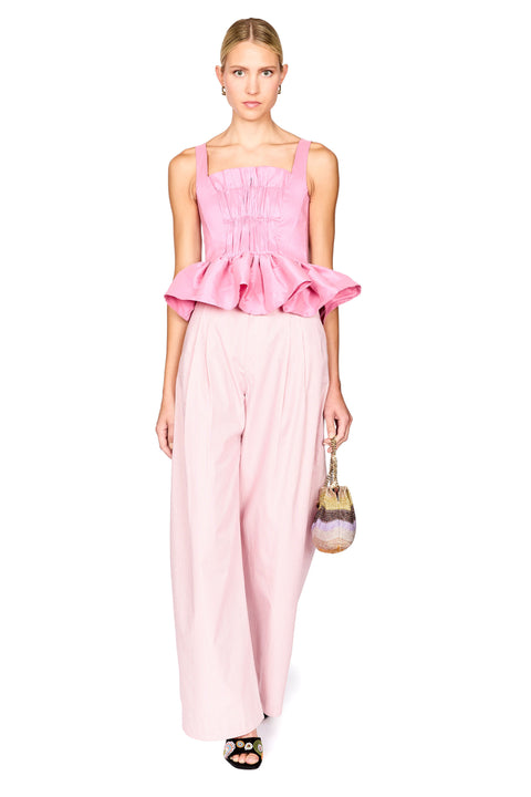 A model wearing a pink sleeveless top with a gathered peplum hem and matching wide-leg trousers, holding a small striped handbag against a plain white background.