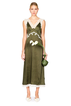 A woman models a sleeveless olive green satin maxi dress with white lace trim at the V-neckline, waist, and hem. She is holding a small green handbag and wearing white pointed-toe flats, set against a plain white studio background.