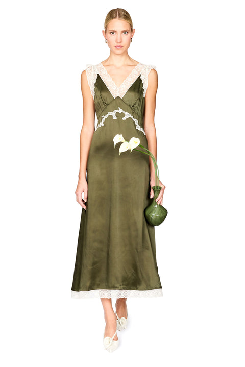 A woman modeling a sleeveless olive green satin slip dress with white lace trim at the neckline, waist, and hem, holding a small green handbag against a plain white background.