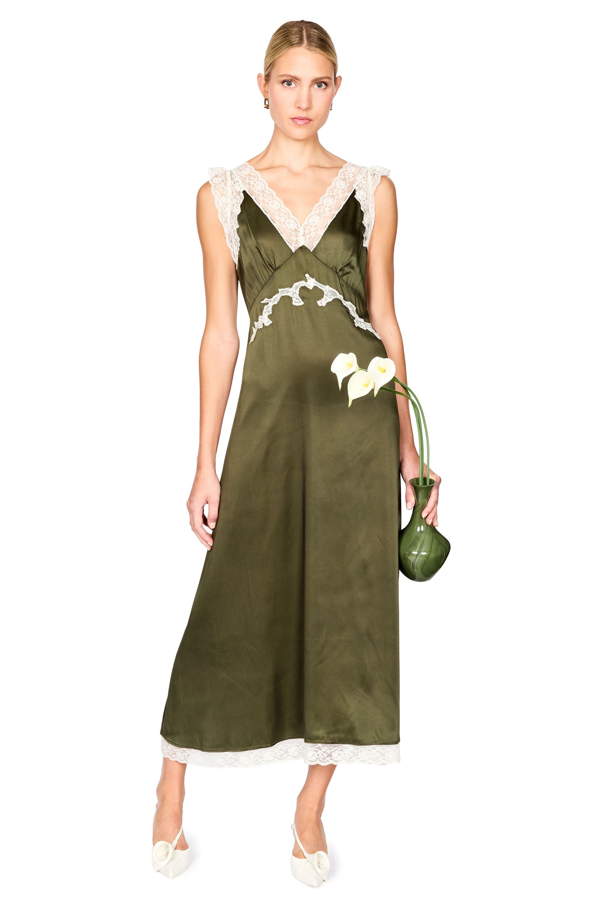 A woman models a sleeveless olive green satin slip dress with white lace trim at the V-neckline and hem, holding a small green clutch vase with white flowers against a plain white background.