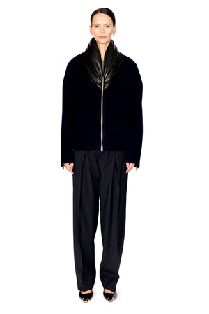A model stands against a solid white background wearing a black velvet jacket with a glossy black shawl collar and a gold front zipper, paired with black wide-leg trousers and black pointed-toe heels with gold detailing.