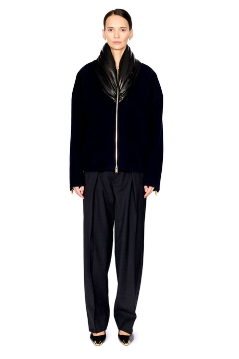 A model stands against a solid white background wearing a black velvet jacket with a glossy black shawl collar and a gold front zipper, paired with black wide-leg trousers and black pointed-toe heels with gold detailing.
