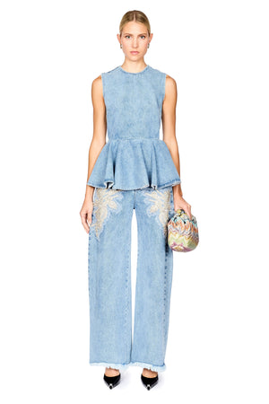A model stands against a white background wearing a light blue denim two-piece set consisting of a sleeveless peplum top and matching wide-leg pants with embroidered details. She is holding a small, colorful patterned clutch and wearing black pointed-toe heels.