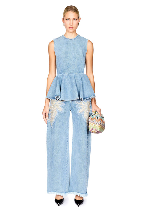 A model stands against a white background wearing a light blue denim two-piece set consisting of a sleeveless peplum top and matching wide-leg pants with embroidered details. She is holding a small, colorful patterned clutch and wearing black pointed-toe heels.