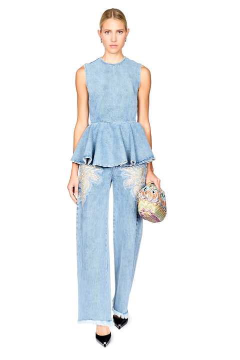 A model stands against a white background wearing a light blue denim two-piece set consisting of a sleeveless peplum top and matching wide-leg pants with embroidered details, holding a small multicolored patterned clutch.