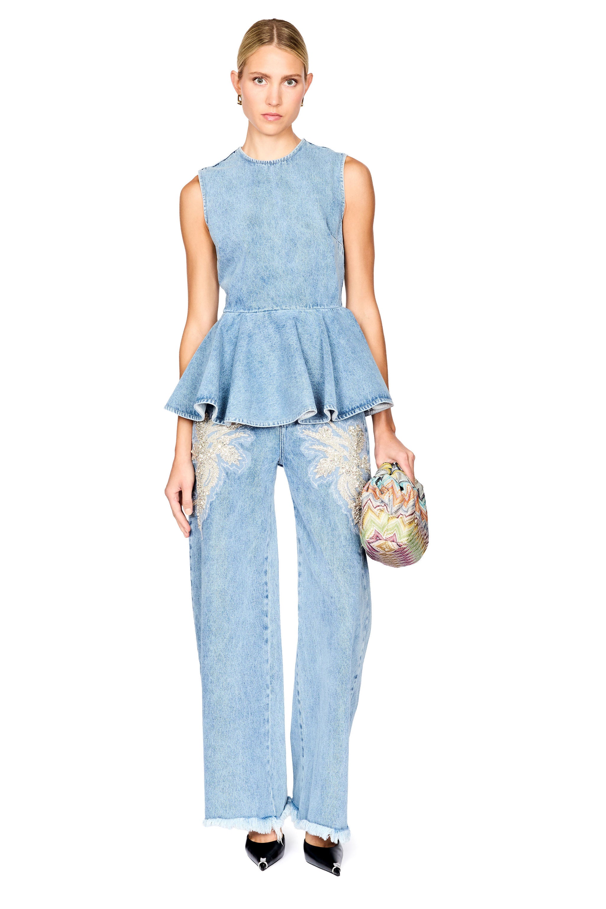 A model stands against a white background wearing a light blue denim two-piece set consisting of a sleeveless peplum top and wide-leg jeans with embroidered details, holding a small patterned clutch.