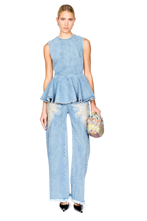 A model stands against a white background wearing a light blue denim two-piece set consisting of a sleeveless peplum top and wide-leg jeans with embroidered details, holding a small patterned clutch.