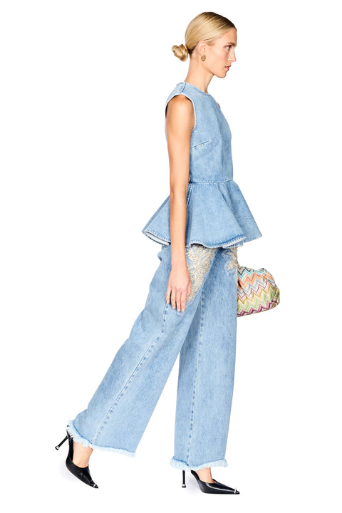 A woman in a light blue denim peplum top and matching wide-leg jeans walks in profile against a white background, holding a small patterned handbag and wearing black pointed heels.