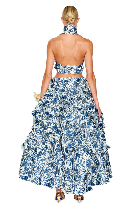 A woman shown from the back wearing a blue and white tropical leaf print halter-neck two-piece set with a ruffled maxi skirt, against a plain white background.