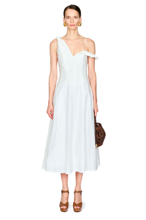 A model wearing a white one-shoulder midi dress with a tie detail at the neckline, paired with brown strappy sandals and holding a brown patterned handbag, set against a plain white background.