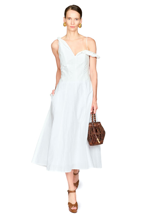 A model wearing a white cold-shoulder midi dress with a V-neckline, holding a brown woven handbag and wearing brown strappy sandals against a plain white background.
