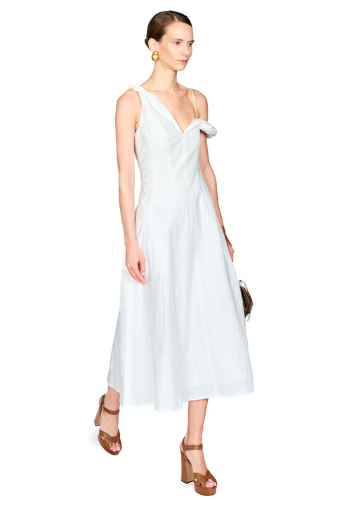 A woman modeling a sleeveless white midi dress with a draped neckline, thin shoulder straps, and a small decorative fabric accent on one shoulder, paired with brown strappy high-heeled sandals against a plain white background.