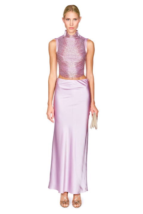A model wearing a two-piece lavender formal set consisting of a sleeveless high-neck textured top and a matching satin skirt, paired with metallic heels and holding a small clutch, on a white background.