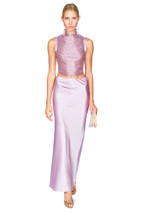 A model wearing a two-piece lavender formal set consisting of a sleeveless, high-neck, textured metallic top and a matching satin maxi skirt, holding a small beige clutch against a white background.