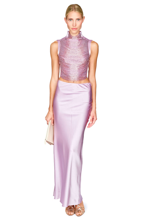 A model wearing a two-piece lavender formal set consisting of a sleeveless, textured high-neck crop top and a matching satin maxi skirt, holding a small beige clutch against a white background.