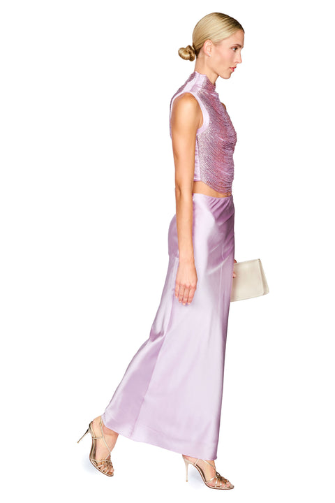 A woman modeling a lavender satin two-piece set consisting of a sleeveless high-neck crop top and a matching wide-leg skirt, paired with nude high heels and a small cream clutch, set against a plain white background.