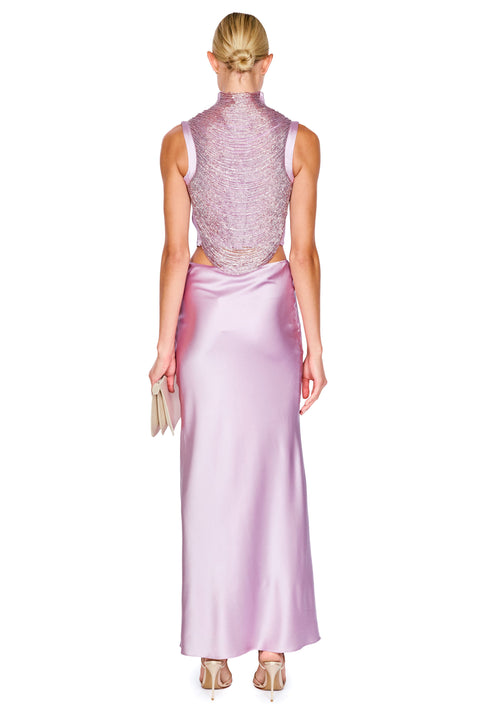 A woman viewed from the back wearing a sleeveless high-neck maxi dress with a heavily embellished beaded lavender-pink back and a smooth satin-like light purple skirt. She is holding a small metallic clutch against a plain white background.