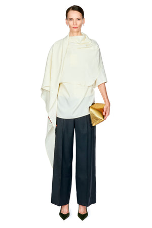 A model stands against a white background wearing a cream draped top, dark black wide-leg trousers, and holding a gold textured clutch.