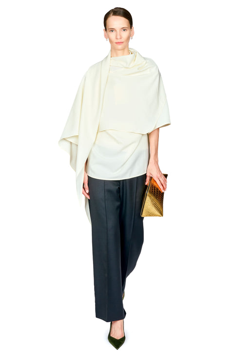A model wearing a cream draped cape top, dark black wide-leg trousers, and olive green pointed shoes, holding a gold textured clutch against a plain white background.
