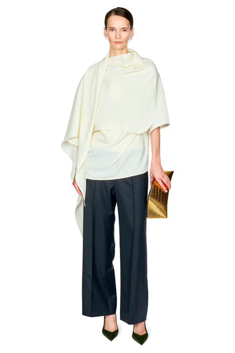 A model stands against a white background wearing a cream draped cape-style top, dark black wide-leg trousers, olive green pointed heels, and holding a gold textured clutch.