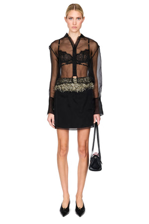 A model stands against a white studio background wearing a sheer black long-sleeve top over a black lace bralette, a black mini skirt with a wide embellished gold waistband, and black pointed shoes, while holding a small black handbag.