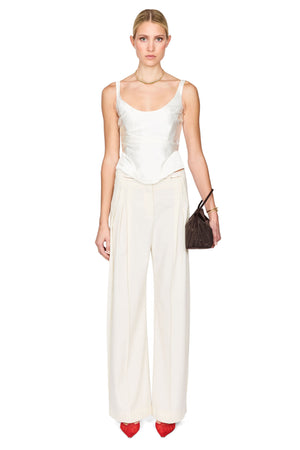 A model wearing a white sleeveless camisole top and matching white wide-leg trousers, styled with red pointed-toe heels and a dark brown handbag, against a plain white background.