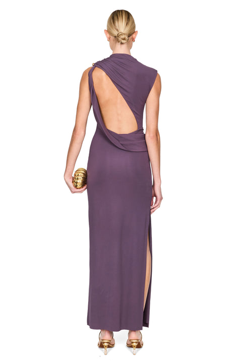 A woman shown from the back wearing a sleeveless, floor-length mauve dress with a dramatic open back and high side slit. She is holding a small gold clutch against a plain white studio background.