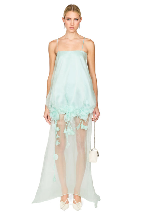 A model wearing a light mint green sheer maxi dress with spaghetti straps, a voluminous gathered skirt with tassel details, and holding a small white handbag against a solid white background.