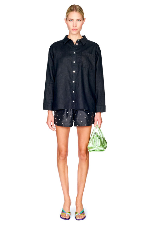 A model stands against a white background wearing a black long-sleeve button-up shirt, black shorts with white polka dots, and holding a green patterned handbag.