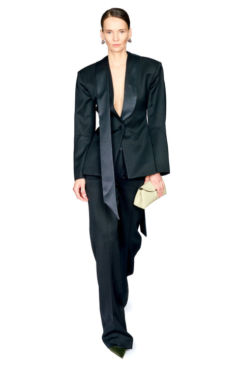 A model wearing a black satin-like tailored suit with a dramatic asymmetrical draped sash, holding a small beige clutch against a plain white background.