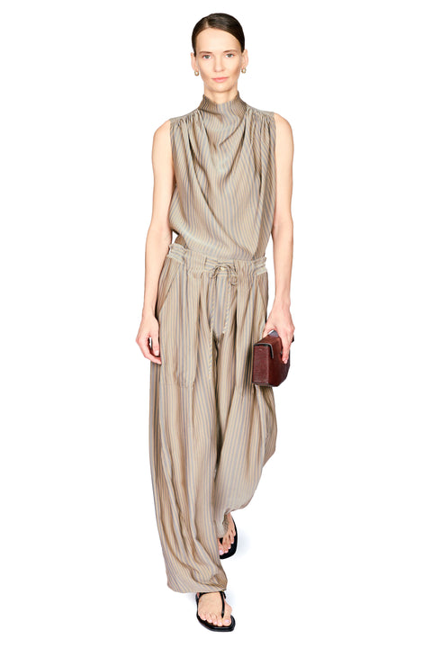 A model wearing a sleeveless, high-neck beige top with vertical pinstripes, and pants with wide draped legs, and a matching tie belt, holding a small dark brown leather clutch against a plain white background.