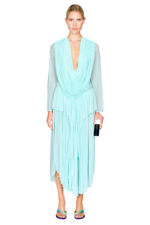 A model wearing a light aqua blue pleated long-sleeve top and maxi skirt set with a draped V-neckline, paired with colorful flat sandals and holding a black clutch, set against a plain white background.