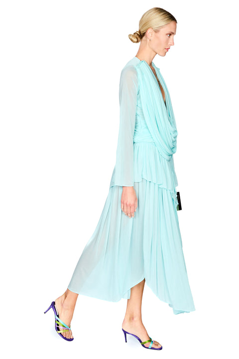 A woman modeling a light aqua blue draped maxi dress with sheer long sleeves and a high-low hemline, paired with purple heeled sandals, against a plain white background.