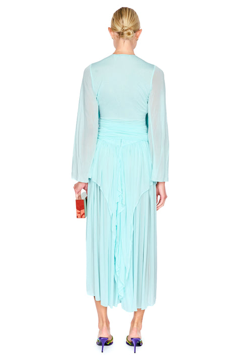 A model wearing a light aqua blue pleated long-sleeve top and maxi skirt set with a draped V-neckline, paired with colorful flat sandals and holding a black clutch, set against a plain white background.