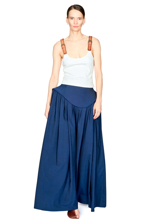 A model wearing a white tank top with brown shoulder straps and a long, flowing navy blue skirt or poncho-style bottom, standing against a plain white background.