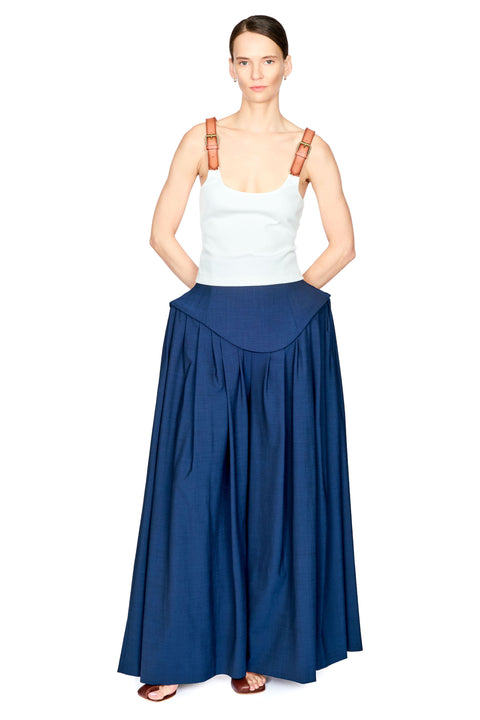A model wearing a long navy blue pleated skirt with a curved waistband, styled with a white tank top and brown shoulder straps, on a solid white background.