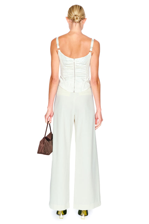 A woman shown from the back wearing a white sleeveless top and pants set with thin shoulder straps, a ruched back, and wide-leg pants. She is holding a small brown handbag against a plain white studio background.