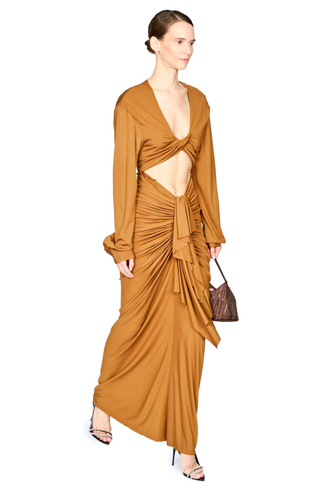 A model wearing a long-sleeved, rust-brown draped maxi dress with a twisted front detail and side cutout, holding a small brown handbag against a plain white background.